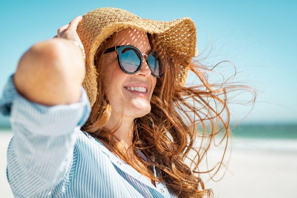 Nainen suojaa ihonsa hatulla ja aurinkolaseilla rannalla.